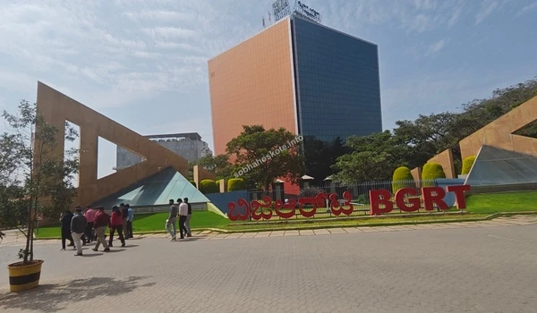 Side view of the large red BGRT lawn signage with the modern glass building facade in the background near Sobha Hoskote